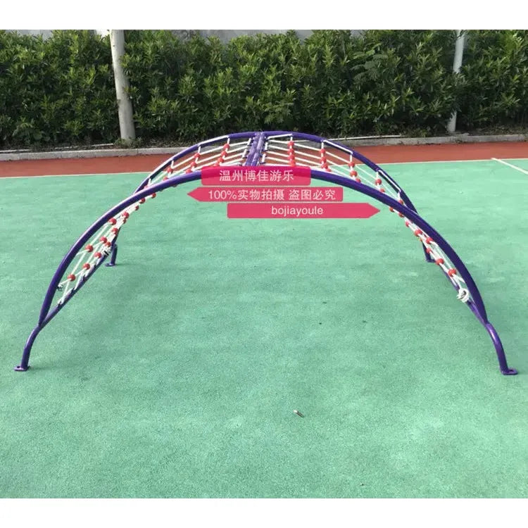 A unisex purple climbing frame with a woven design stretches across the playground, suitable for ages 3-8. This vibrant metal addition is complemented by a red sign and lush green bushes, enhancing its serene backdrop. Product: outdoor toys Childrens swings Climbing nets Sensory integration Sports equipment.