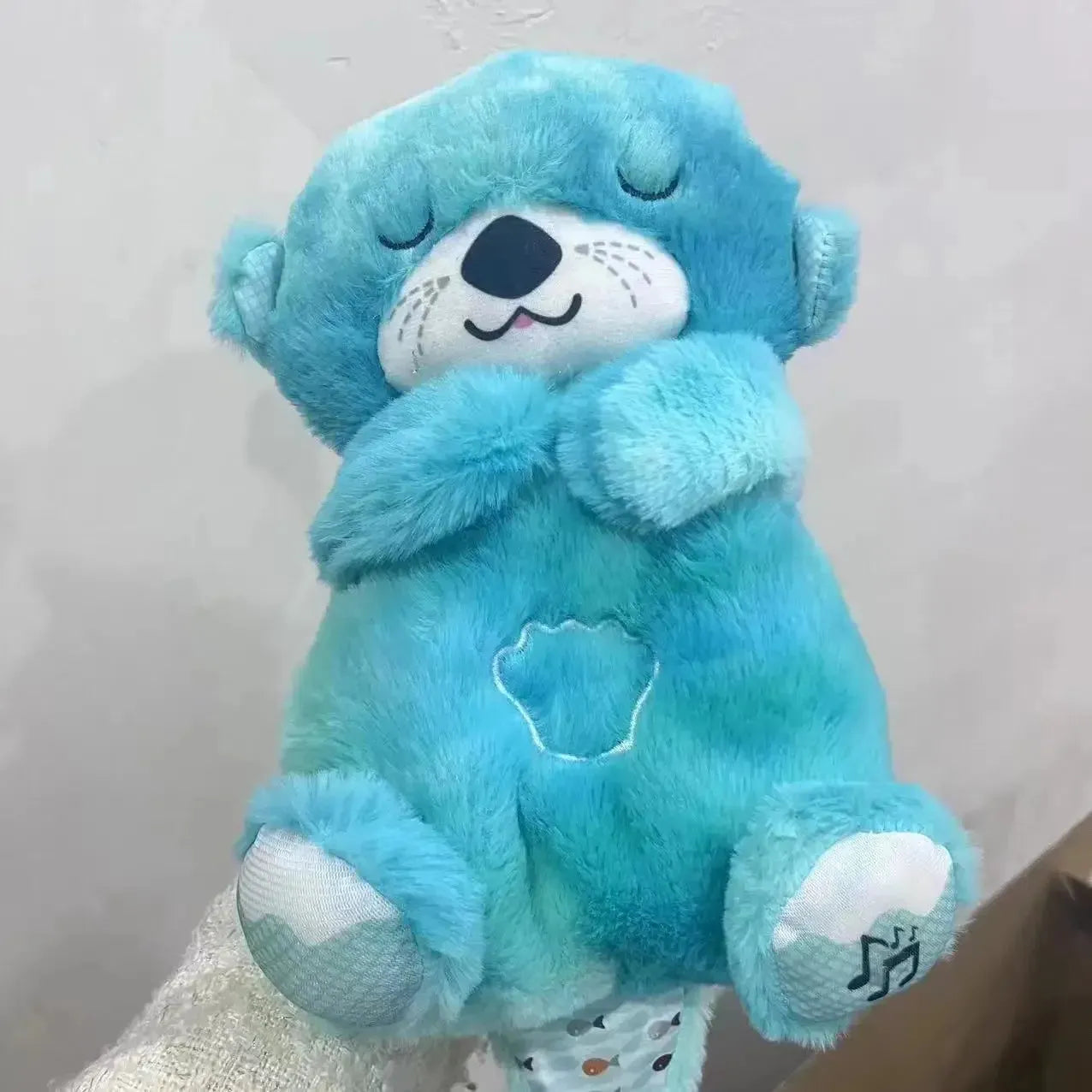 The Soothing Otter Musical Plush Toy, designed for newborns, closes its eyes contentedly with crossed arms. It features a heart-shaped belly patch and a musical note on a white paw, offering a comforting sensory sleep experience.