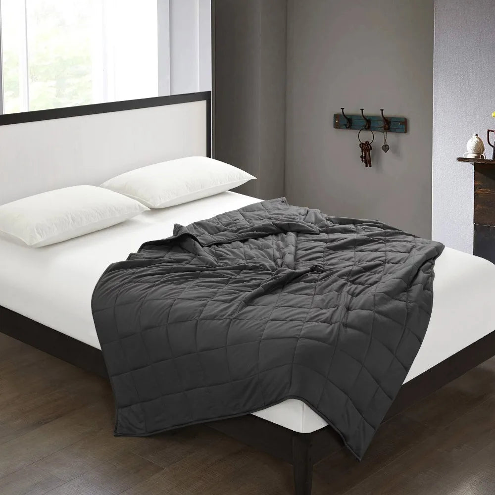 A neatly made bed with a white sheet and pillows showcases the Plush 20lb Weighted Blanket, expertly crafted for relaxation. The rooms hardwood floors, light gray walls, and a sunlit window enhance the ambiance, while a hook with keys is visible by a small mantle on the wall.