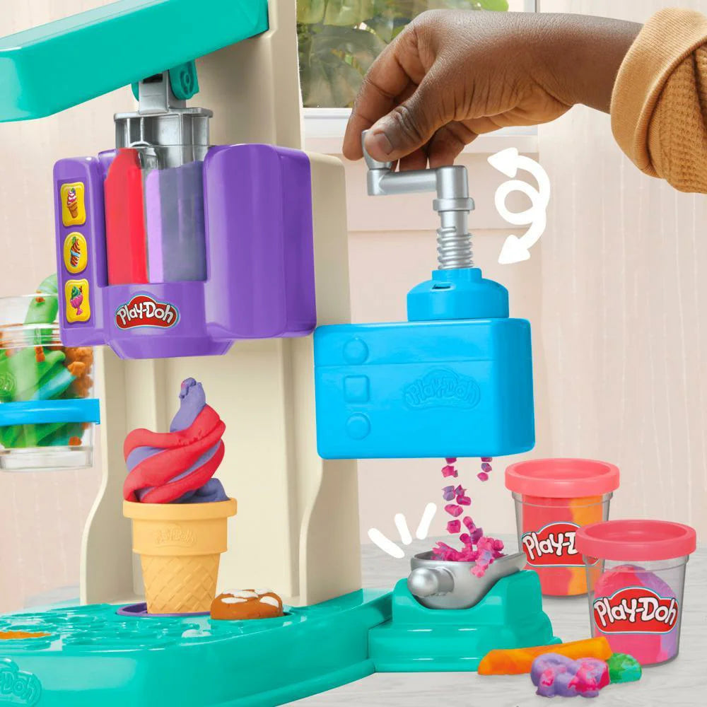 A child expertly uses the Hasbro Play Doh Rainbow Swirl Ice Cream Playset, pulling a lever to swirl pink and purple dough into a cone, with vibrant tubs of Play-Doh adding to the imaginative and playful ambiance in the background.