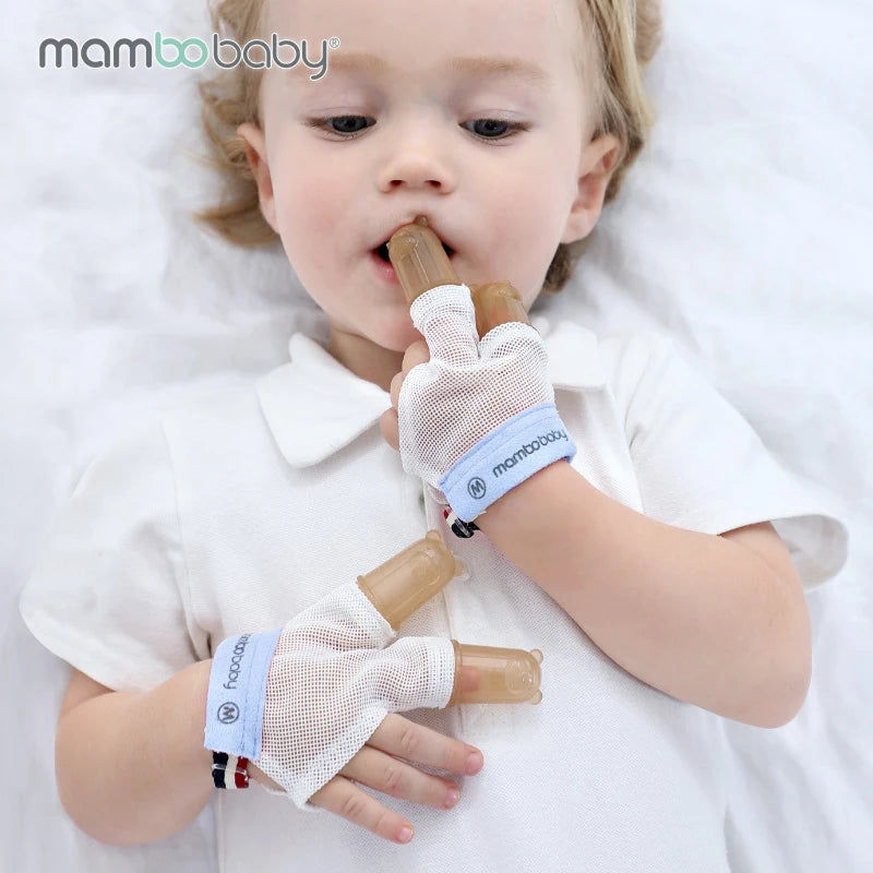A toddler in a white shirt, lying down and soothing teething discomfort, wears Mambobaby Anti-Nail Biting Gloves with brown silicone finger cots on three fingers to manage thumb sucking.