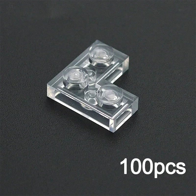 A transparent L-shaped plastic building block from the MOC Transparent Bulk Brick Set (L Transparent), featuring three studs, rests on a black surface. Compatible with Lego. 100pcs appears in white at the lower right corner.