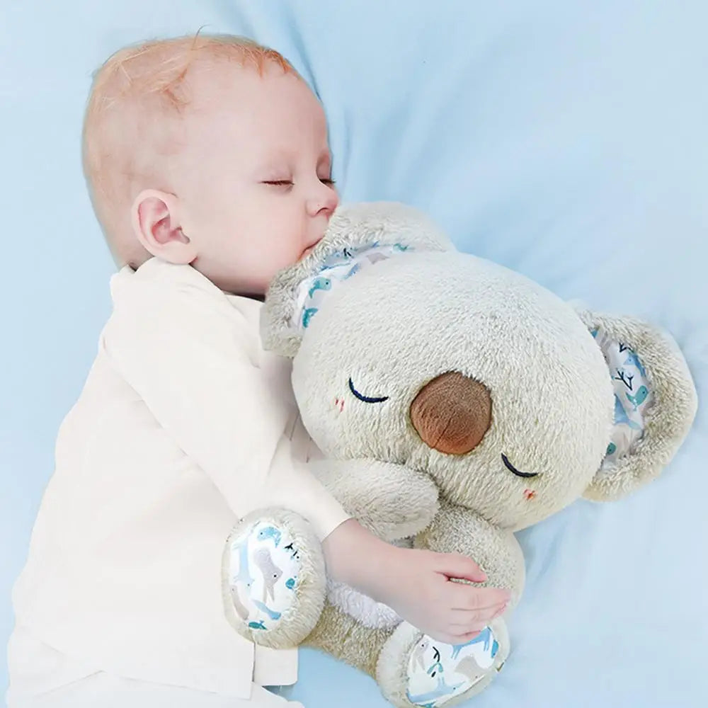A baby in a white outfit sleeps peacefully on a light blue surface, cuddling a Koala Night Light Plush Toy: Musical Sensory Sleep Aid for Newborns, which hums soothing melodies. The koala features closed eyes, blush marks, and patterned ears and feet.