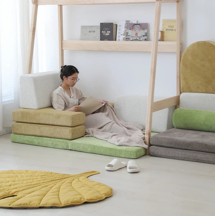 A person relaxes on cushions, absorbed in a book. The minimalist room has a sleek shelf for books, a yellow leaf-shaped rug, and slippers. A Versatile Solid Wood Childrens Bed with Climbing Frame and Swing Features adds functionality, while neutral tones create coziness with natural textures.