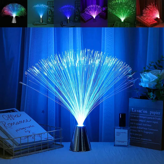 The Multicolor LED Fiber Optic Night Light, featuring a metallic base and glowing blue strands, sits beside a decorative basket with books and bottles. The lamps purple, green, and red hues resemble a starry sky light, complemented by an elegant white flower arrangement.