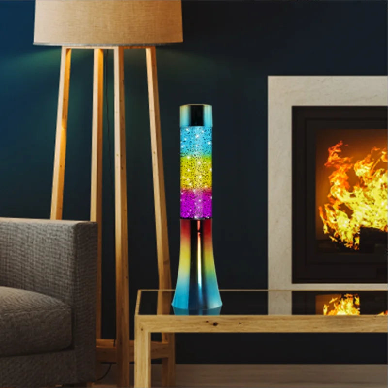 A cozy living room with a lit fireplace and modern floor lamp features the Color-Changing LED Glitter Lava Lamp - 13 Inch Novelty Night Light, adding sparkle on a glass-top table. The gray upholstered sofa enhances the warm ambiance with its perfect decorative elements.