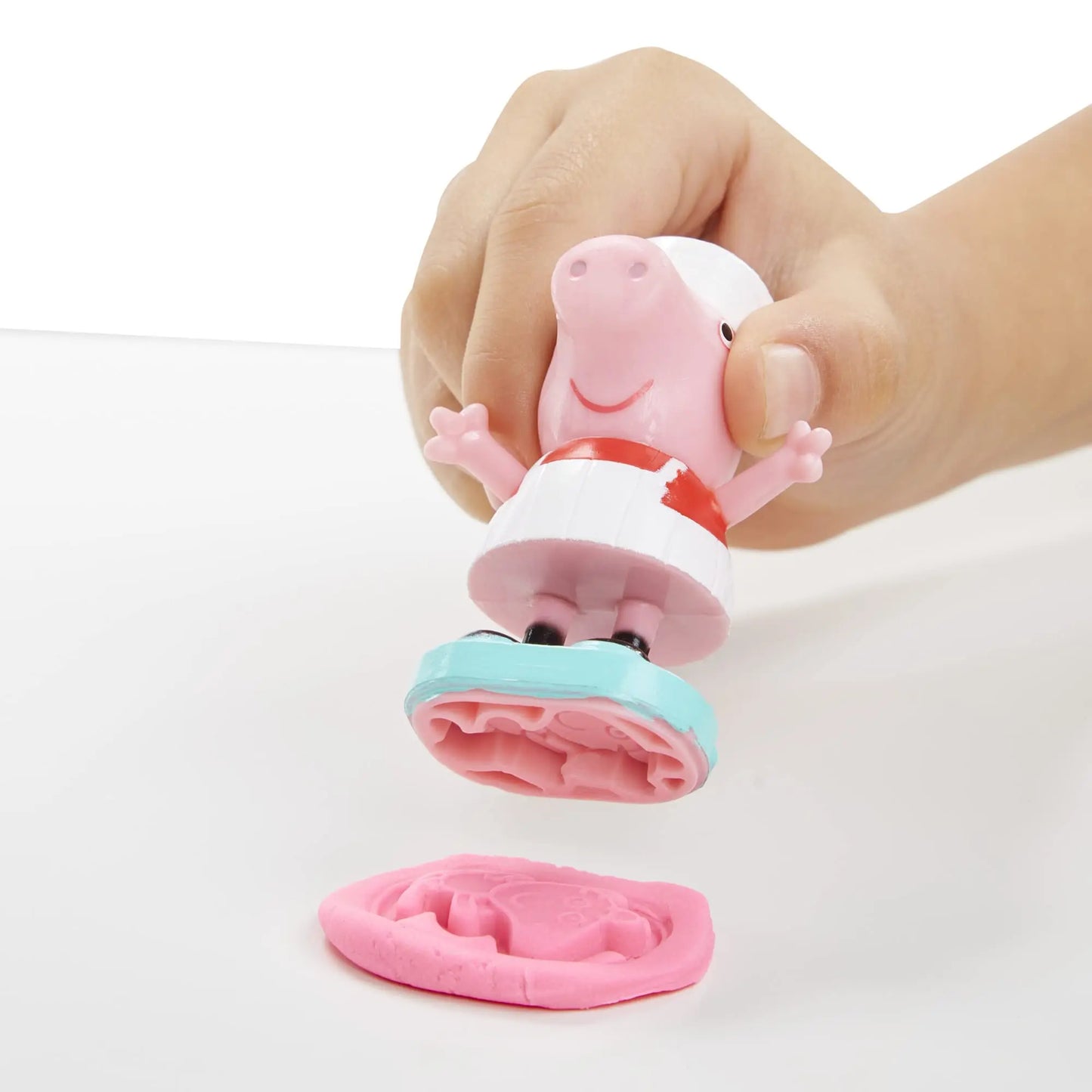 A hand uses a stamp from the Play-Doh Peppas Ice Cream Playset with a toy shaped like Peppa Pig, dressed in white and red, to imprint her joyful outline onto pink modeling clay.