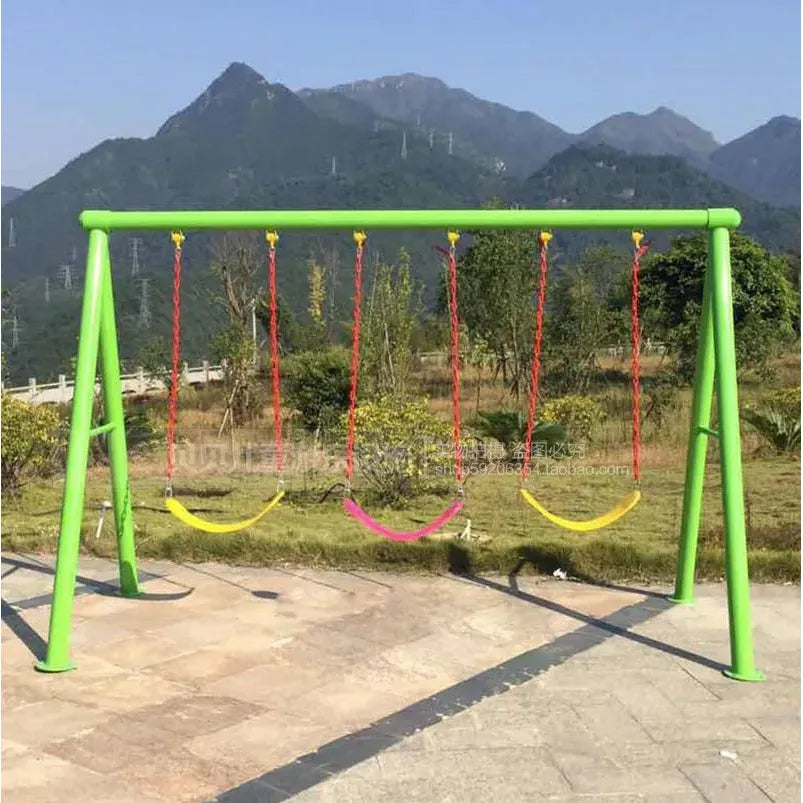 A green Outdoor Steel Climbing Frame Swing features four colorful swings—three yellow, one pink. Set against green mountains and a clear blue sky, it creates an ideal environment on this inviting paved area for childrens sensory integration training.