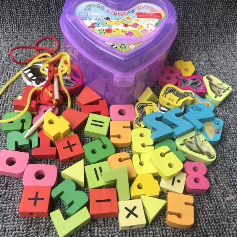 A vibrant assortment of Baby Wooden Lacing Beads, featuring numbers, shapes, and math symbols, is spread on a gray surface. A purple heart-shaped box sits behind them with laces to the left for threading. This 50-piece set is ideal for educational play and enhancing fine motor skills.