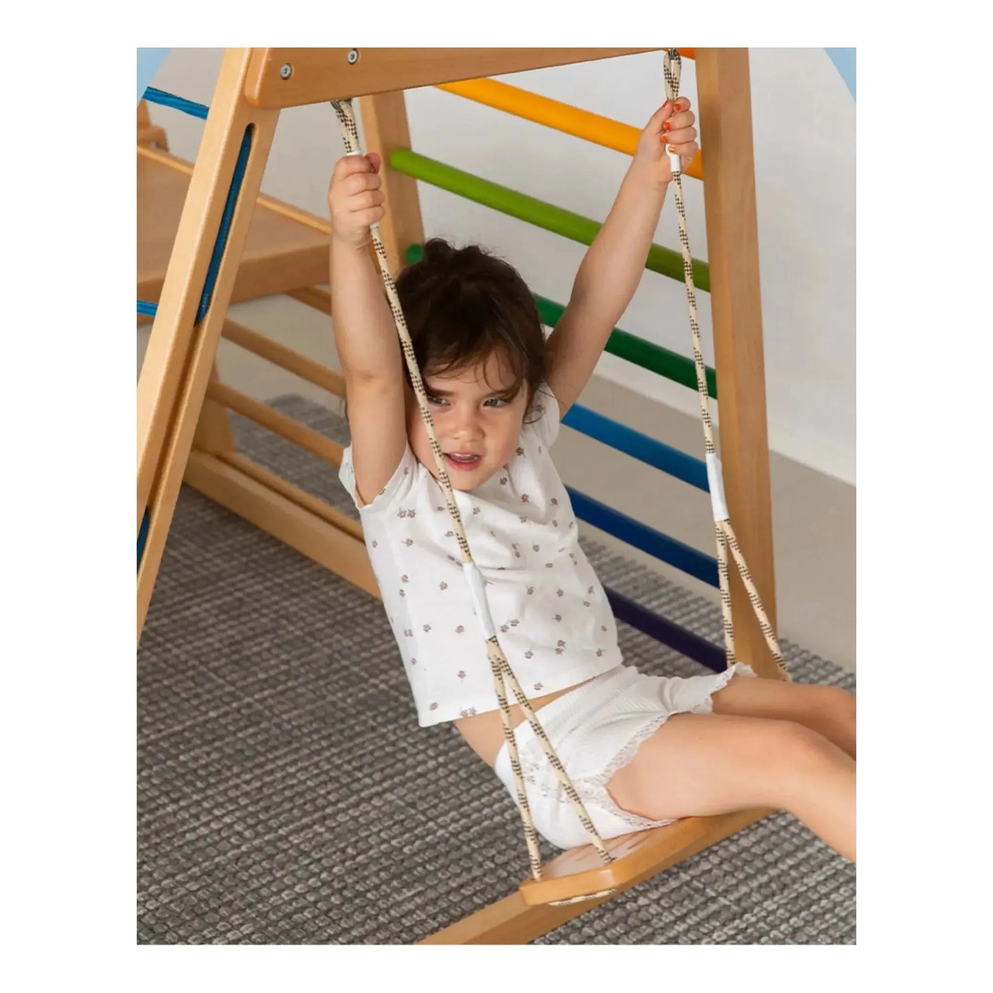 A young child, in a white outfit, joyfully swings indoors on the Indoor Adventure Climbing Frame for Kids with Slide and Swing - Family Fun Playground. Colorful bars and a textured rug enhance the background as they smile and cling to the ropes, delighting in this playful escape.