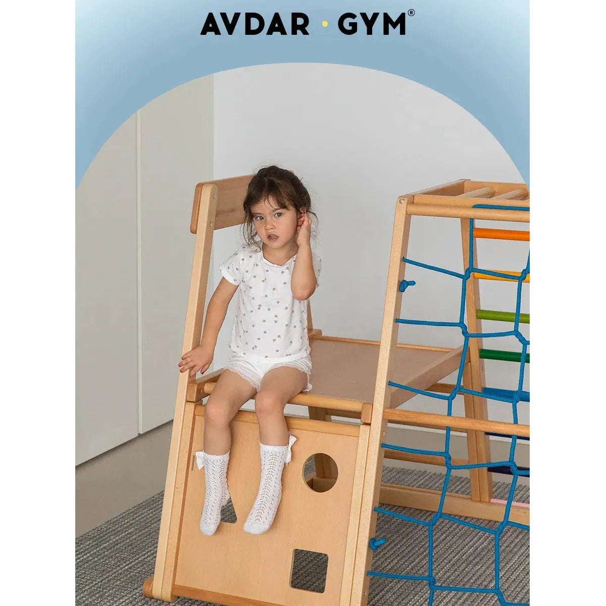 A young child in a polka-dot shirt and shorts sits on a wooden climbing frame with a blue net. The bright, neutral interior features an AVDAR GYM sign. The product is the Indoor Adventure Climbing Frame for Kids with Slide and Swing – Family Fun Playground.
