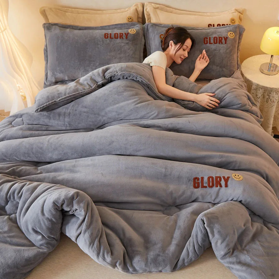 A woman lounges on a bed under the KDJEIKZ Ultra Cozy Winter Weighted Blanket, featuring luxurious milk velvet and adorned with GLORY and a smiley face. Fluffy gray bedding complements the ensemble, while a lamp sits on the bedside table.