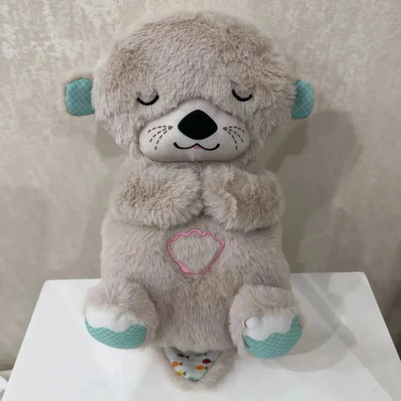 A plush gray otter baby toy, featuring light blue ears and paws, closed eyes, and a stitched pink seashell on its belly, known as the Soothing Otter Musical Plush Toy with Light and Sound for Newborns, sits on a white surface against a beige background.