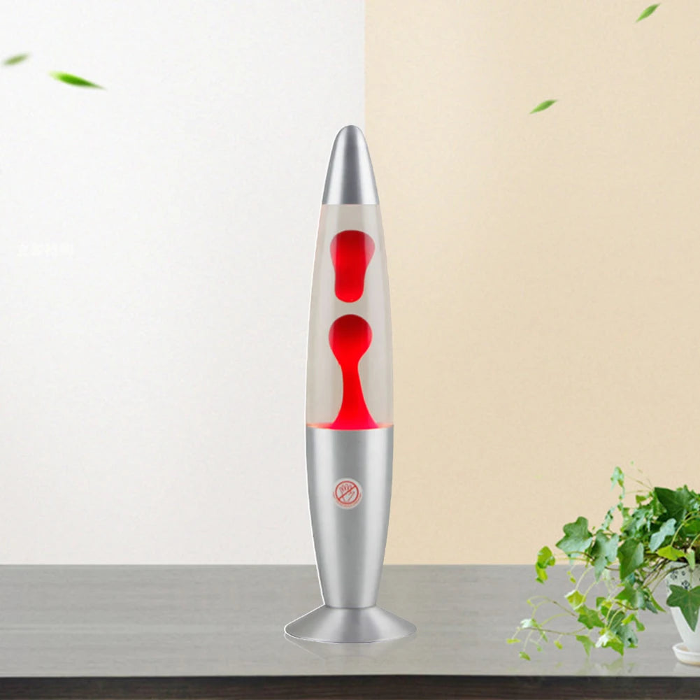 A Luminous Volcano Lava Jellyfish LED Night Light, with a silver hue and red wax, rests on a gray surface. The ambiance is relaxing, set against a background split into beige and light brown. Green leaves float as a leafy plant decorates the bottom right corner, enhancing tranquility.