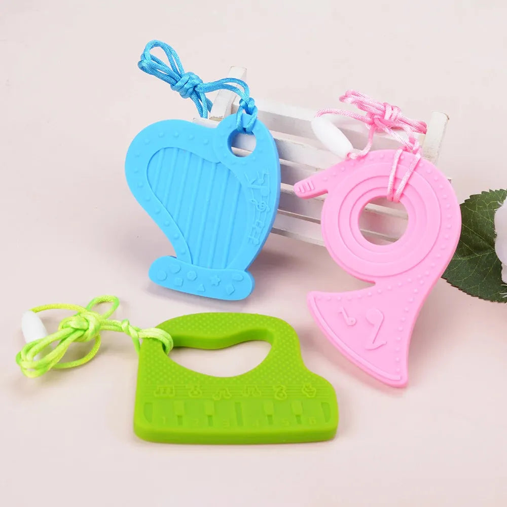 Three vibrant silicone teething toys lay on a light surface: a blue guitar, pink horse, and green rectangle with textures and strings. A ladder and green leaves enhance the scenes charm. These Silicone Chewable Necklaces for Kids are ideal as soothing sensory pendants for autism and ADHD therapy.