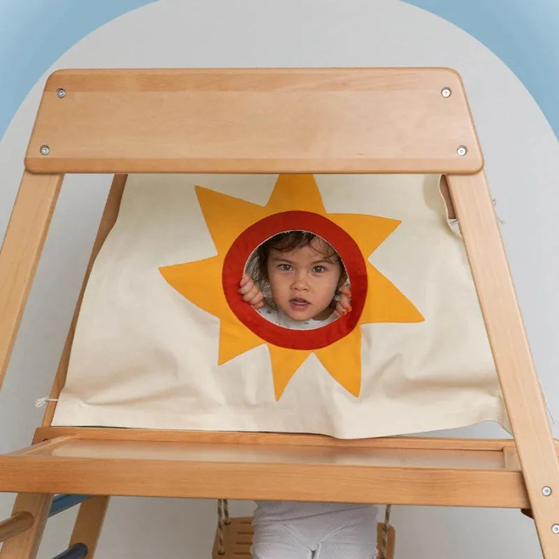 A child peeks through a sun-decorated opening in the Indoor Adventure Climbing Frame for Kids with Slide and Swing. This wooden structure, part of an engaging playground, features a soft blue and white color scheme that adds to its inviting charm.