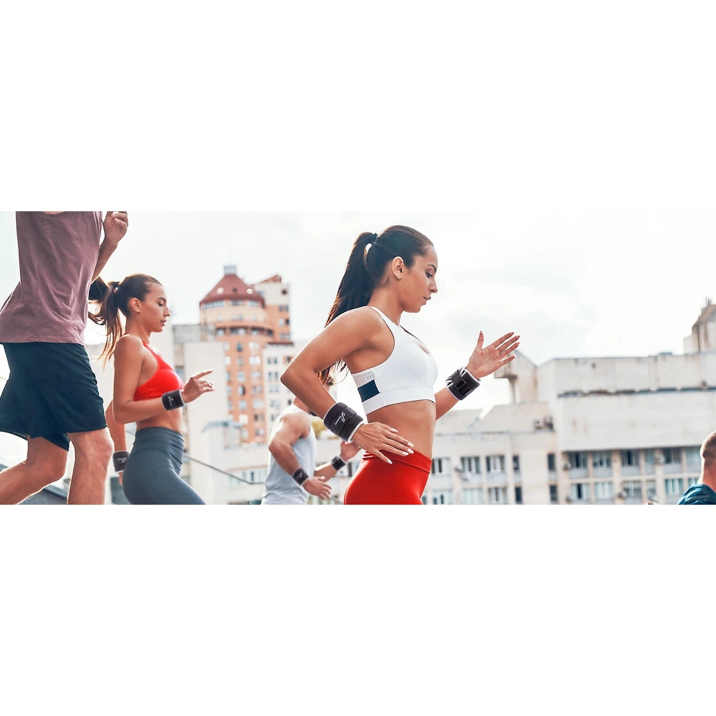 A group jogs outdoors in an urban area, highlighting a woman wearing a white sports bra and red leggings with the Sportneer Adjustable Ankle and Wrist Weights Set for Enhanced Strength Training. Others are in athletic wear, with city buildings and a partly cloudy sky visible in the background.