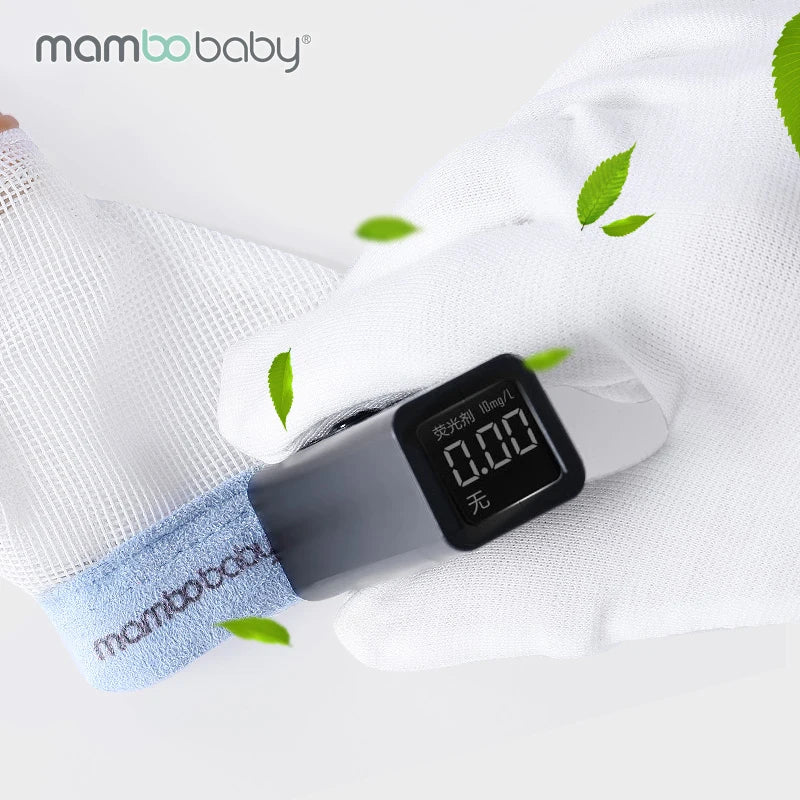 A close-up of a hand in Mambobaby Anti-Nail Biting Gloves with silicone finger cots, holding a digital device with a numeric display. Attached to white and blue fabric labeled mambobaby, it is adorned with green leaves, and the mambobaby logo is at the top left.