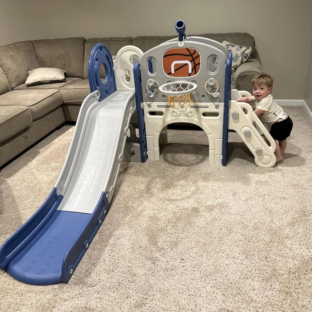 A toddler stands by the 8 In 1 Toddler Slide Set With Basketball Hoop For Kids, set up on a carpeted living room floor with a sectional sofa in the background.