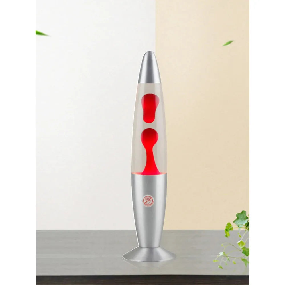 The Luminous Volcano Lava Jellyfish LED Night Light in metallic gray, casting red lava blobs, creates a relaxing ambiance on the table. A split wall with cream and beige hues forms the background as green leaves softly encroach from the right and top left corners.