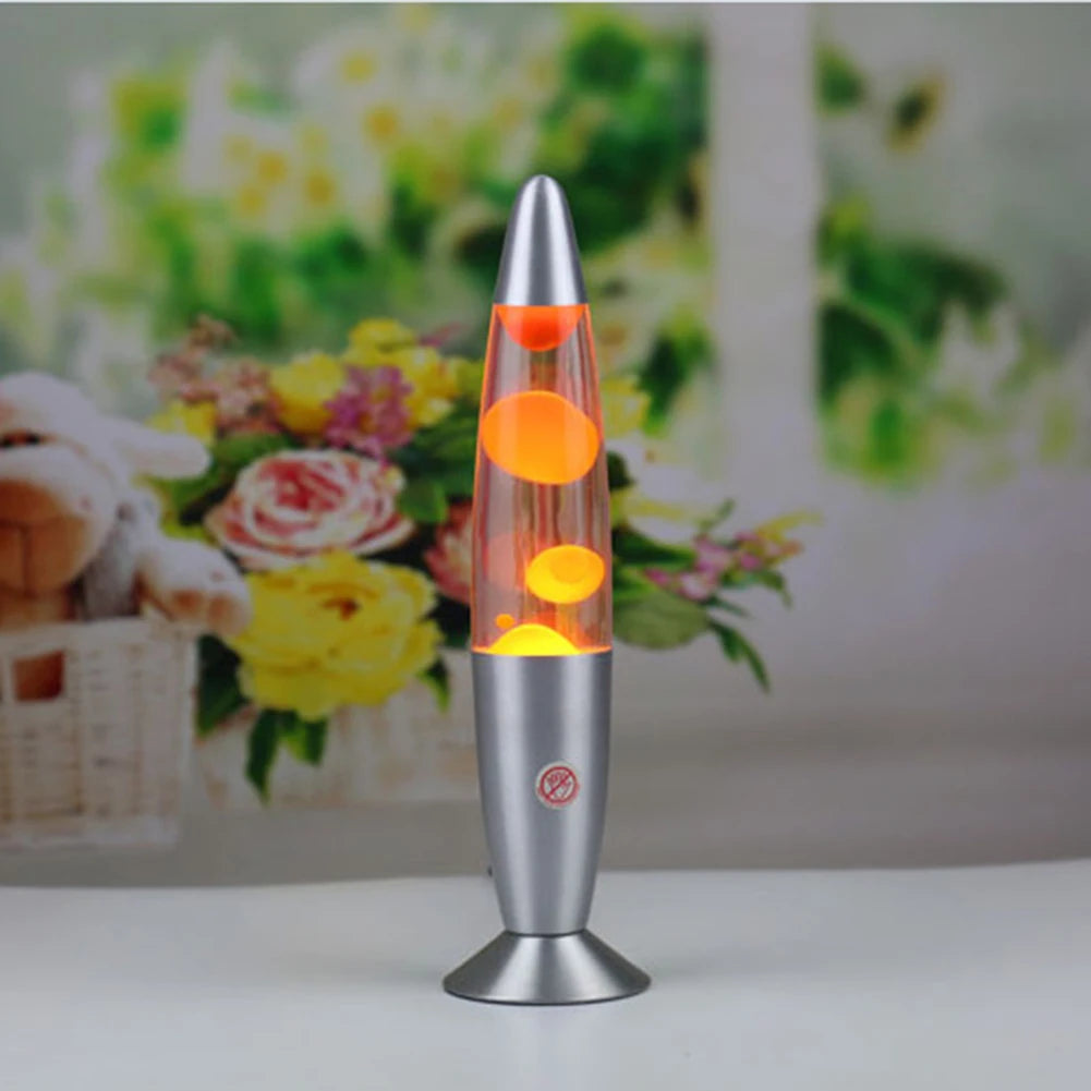 The Luminous Volcano Lava Jellyfish LED Night Light, a creative 25W atmosphere lamp, offers a relaxing ambiance. Its silver base and vibrant orange wax complement any space beautifully, set against a blurred background of yellow and pink blooms.