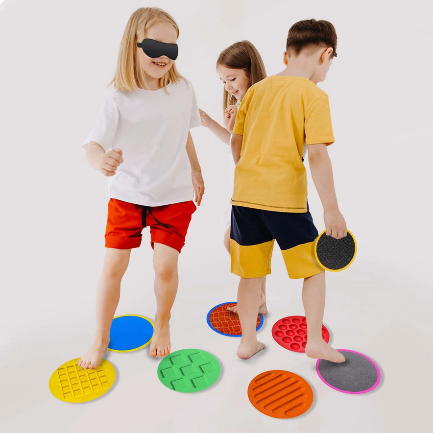 Three children wearing casual clothes engage in developmental play on a colorful Montessori Tactile Adventure Mat with Balance Stones. One child is blindfolded, stepping barefoot on the mat against a plain white background, highlighting their playful exploration.