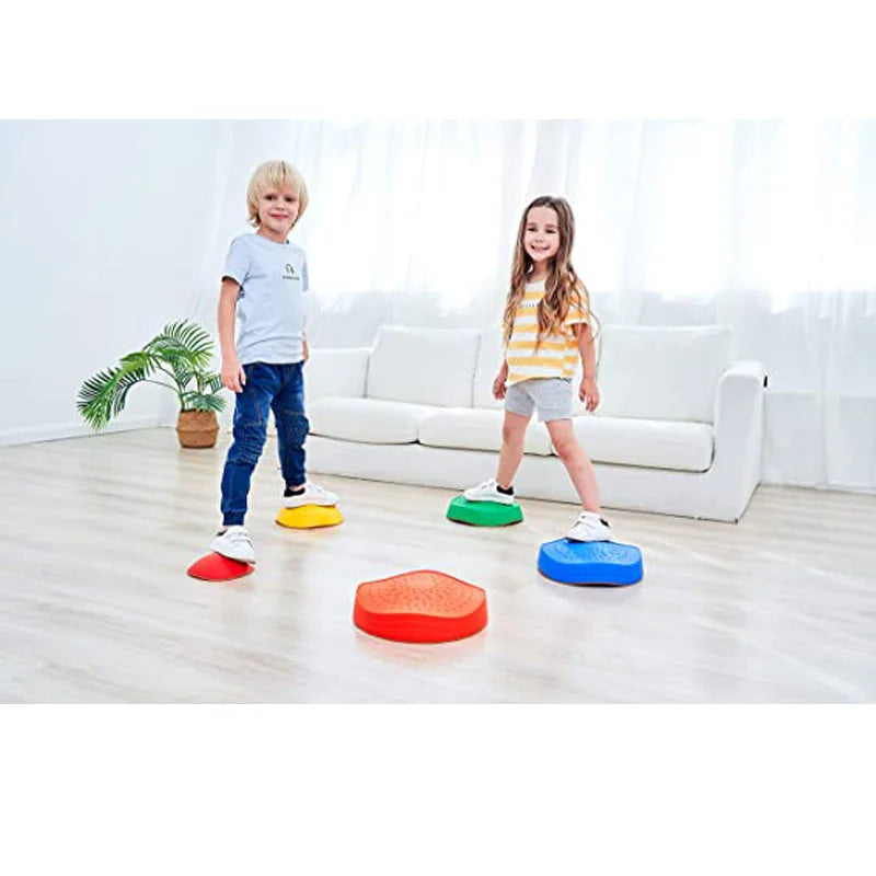 Two children smile on vibrant stepping stones from the Kids 5-Piece Outdoor Balance Stepping Stones Set in a bright living room with a white sofa and plant. The boy wears a white shirt and jeans; the girl, a striped shirt and shorts.