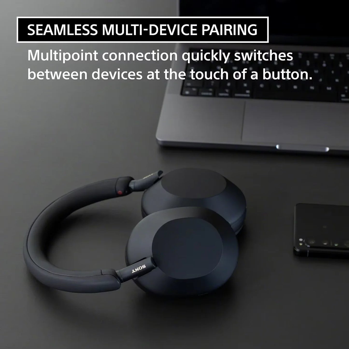 A pair of black Sony WH-1000XM5 Wireless Noise Canceling Headphones with Integrated Processor V1 and Soft Fit Leather rests on a dark desk by a laptop and smartphone. Enjoy seamless multi-device pairing and best-in-class noise cancellation.