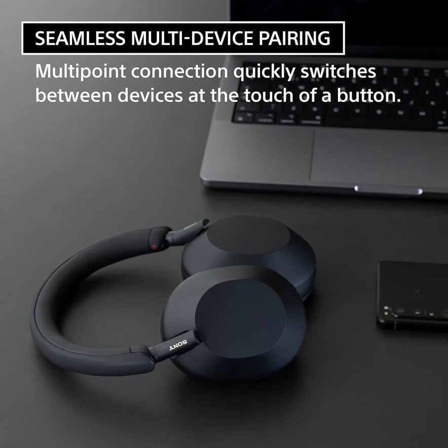 A pair of black Sony WH-1000XM5 Wireless Noise Canceling Headphones with Integrated Processor V1 and Soft Fit Leather rests on a dark desk by a laptop and smartphone. Enjoy seamless multi-device pairing and best-in-class noise cancellation.