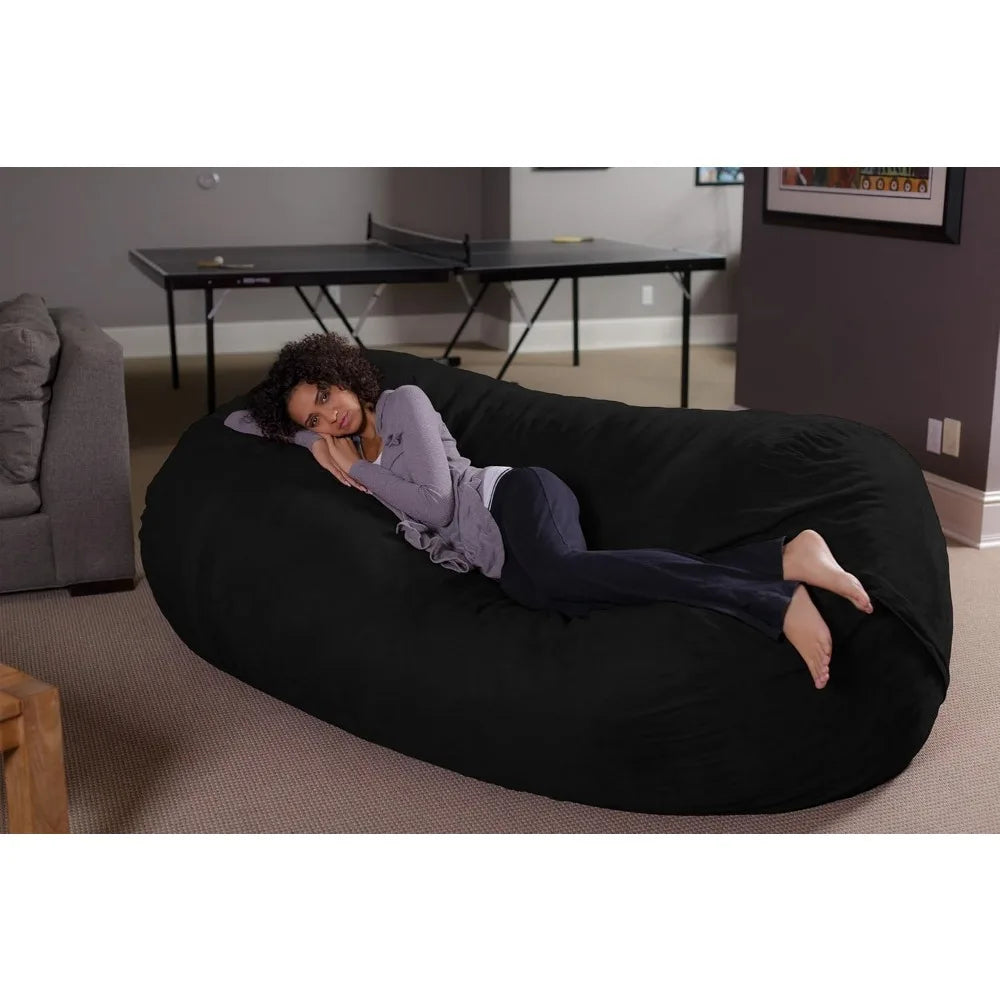 A woman with curly hair relaxes on her side on a Bean Bag Lounger - Plush Sofas with Soft Microsuede Cover in a living room, with a gray sofa and a ping pong table visible in the background.