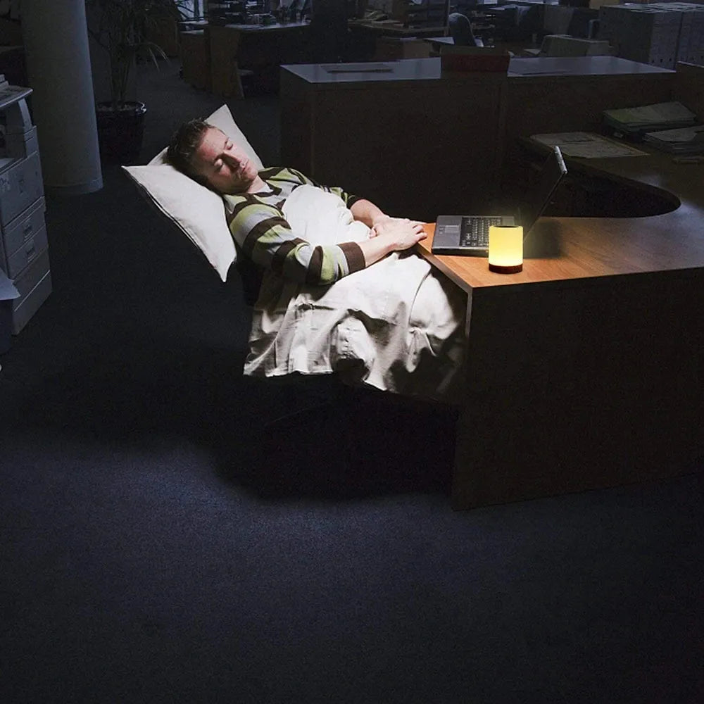 A person sleeps in an office chair, covered by a blanket and resting on a pillow, with the warm glow of a Portable Touch-Sensitive LED Night Light illuminating the dim room. An open laptop sits on the desk, creating an office-like atmosphere.