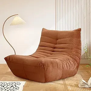 A brown Fireside Chair Bean Bag for adults with a modern, armless design in soft suede sits on a wooden floor beside a curved floor lamp with a pleated shade, complementing the bright minimalist room.