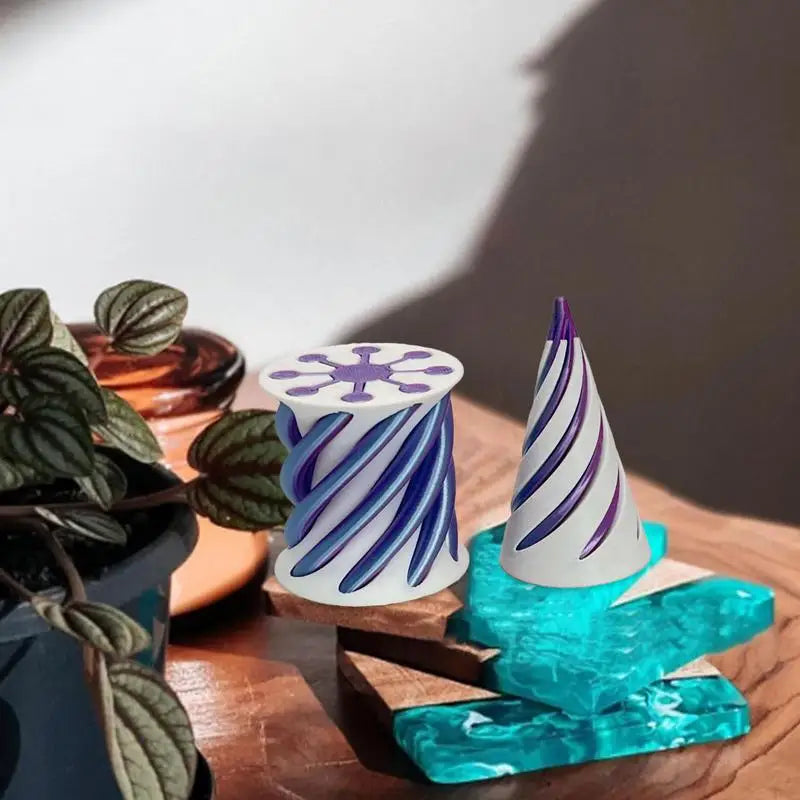 Two small Impossible Cone Sensory Fidgets with white rubber bases and swirling purple patterns—one cylindrical, one cone-shaped—rest on a wooden surface near a potted plant and decorative blue and amber glass pieces.