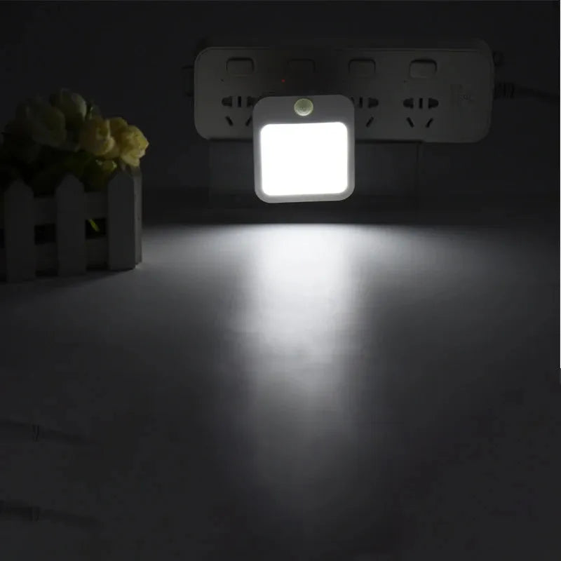 The Dimmable Motion Sensor LED Night Light with EU Plug emits a soft white glow when plugged into an outlet, enhancing the ambiance. Positioned nearby is a decorative piece featuring a small white picket fence and flowers, adding charm to the setting in your babys room or bedroom.