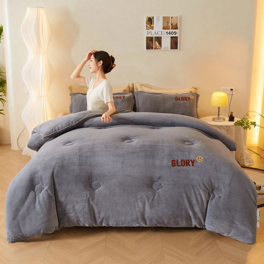 A woman sits on a bed with gray bedding and plush pillows, one boasting GLORY. The room has beige walls and a wooden floor, featuring the KDJEIKZ Ultra Cozy Winter Weighted Blanket for warmth. A tall white lamp stands by a small table adorned with decor behind her.