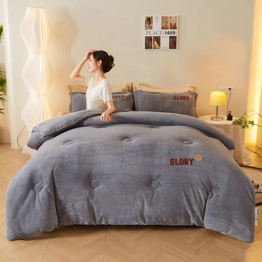 A woman sits on a bed with gray bedding and plush pillows, one boasting GLORY. The room has beige walls and a wooden floor, featuring the KDJEIKZ Ultra Cozy Winter Weighted Blanket for warmth. A tall white lamp stands by a small table adorned with decor behind her.