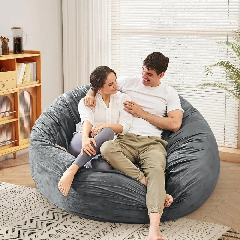 A man and woman smile at each other while sitting on a Bean Bag Chairs Convertible Bean Bag Bed with a microfiber cover in a cozy, sunlit living room. A bookshelf, window blinds, and a potted plant are visible in the background.