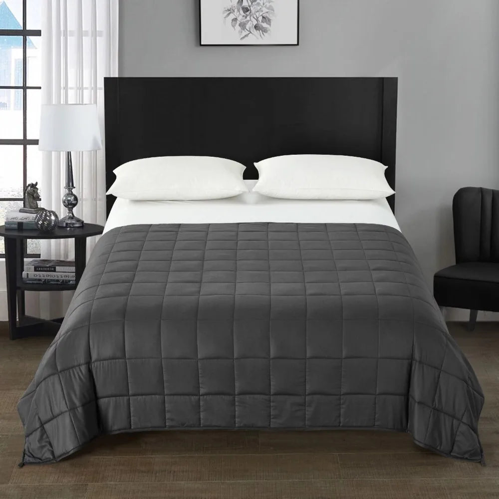 A neatly made bed with a dark headboard is topped with a luxurious plush weighted blanket, showcasing expert material blending. Two white pillows rest elegantly beneath framed black-and-white artwork. A lamp graces the bedside table, and an Origin US chair sits stylishly in the rooms corner.