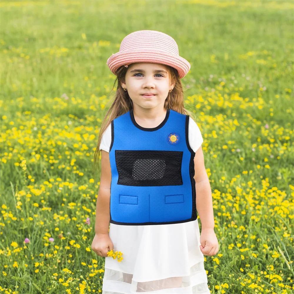 A young girl stands in a field of yellow flowers, wearing a Breathable Adjustable Weight Vest for Kids over a white dress and pink-striped hat. With a serene expression, she looks directly at the camera, surrounded by lush green grass that promotes stress relief.