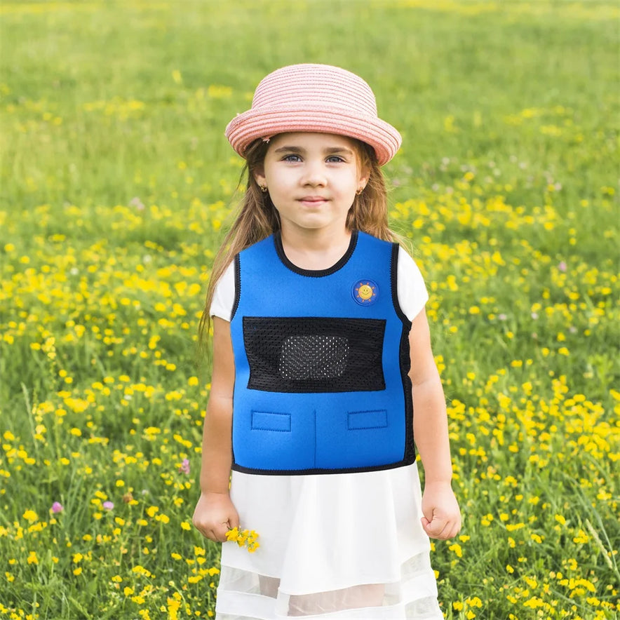 A young girl stands in a field of yellow flowers, wearing a Breathable Adjustable Weight Vest for Kids over a white dress and pink-striped hat. With a serene expression, she looks directly at the camera, surrounded by lush green grass that promotes stress relief.