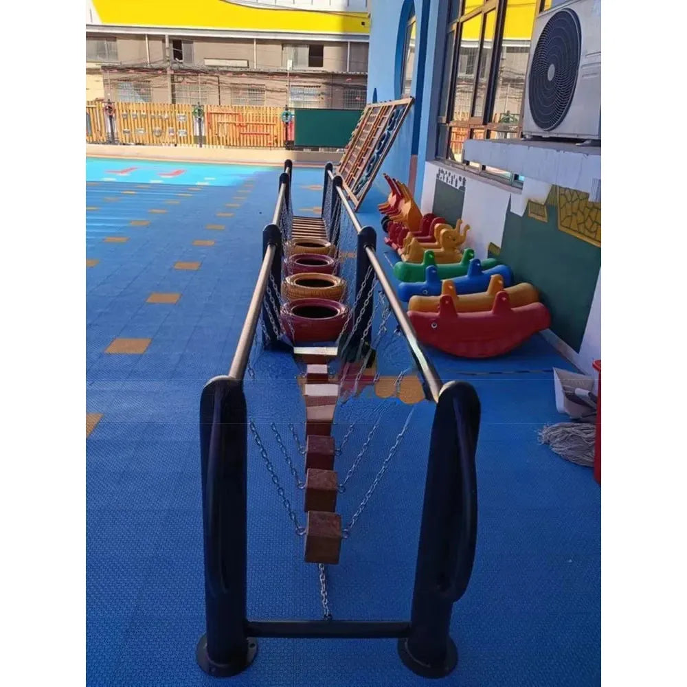 The childrens outdoor play setup includes a rope and block bridge, colorful toy boats, and climbing wall. A yellow structure stands on blue flooring, with an Outdoor Steel Climbing Frame Swing enhancing sensory integration.