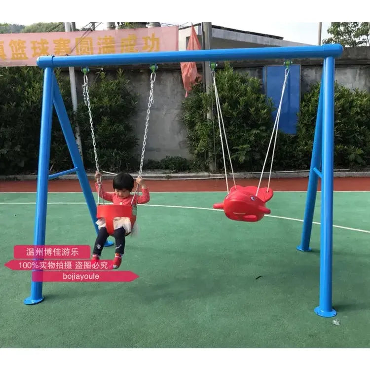 A child in a red shirt is on a swing attached to an outdoor toys Childrens swings set, with a red fish-shaped swing seat beside them. The unisex blue metal set, suitable for ages 3-8, is on a green surface near a track.