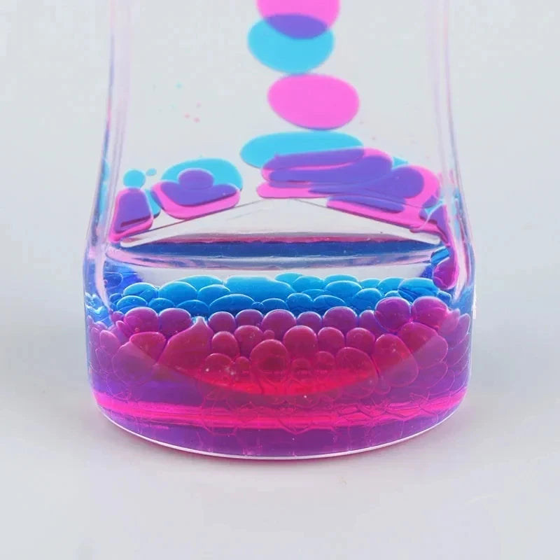 A close-up of the Liquid Motion Bubbler Timer shows pink and blue bubbles in clear liquid inside a transparent container, ideal for sensory activity, anxiety relief, or autism support, set against a light gray background.