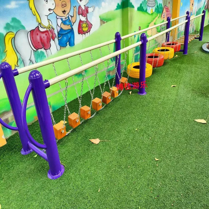 The playground showcases balance beams and chain-suspended stepping blocks, resembling large unisex toys. The area is covered with artificial grass and a colorful mural featuring cartoon characters and rainbows in the background, bordered by vibrant tires.