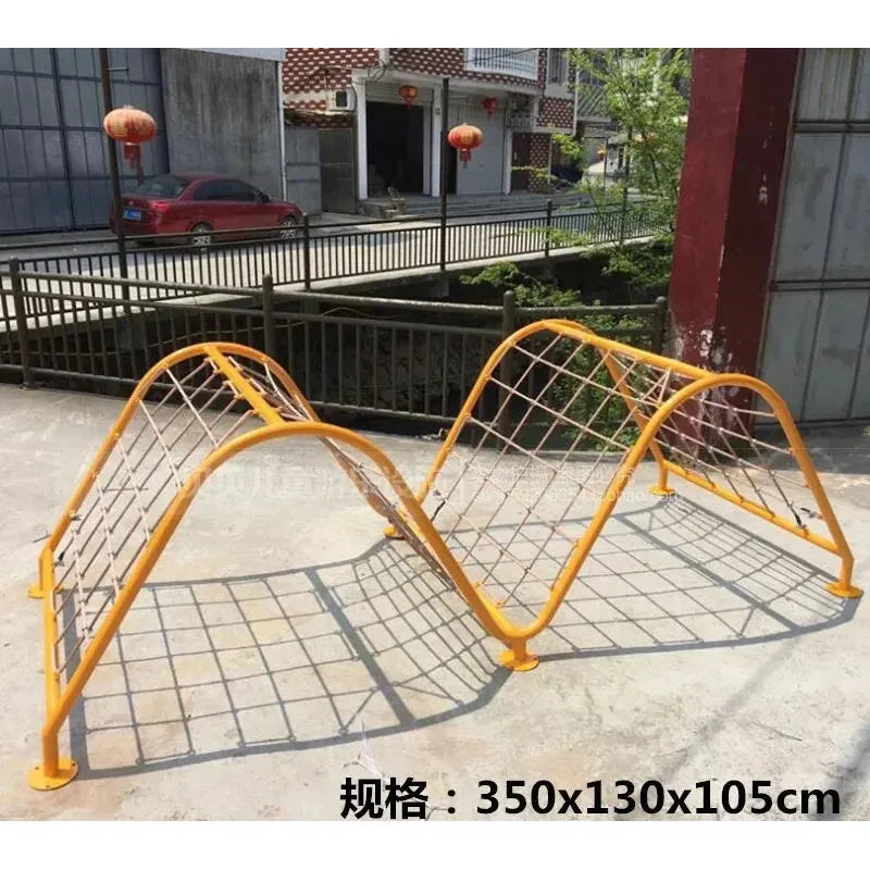 The Outdoor Steel Climbing Frame Swing aids childrens sensory integration with a yellow metal wave-like design and netting. Measuring 350x130x105cm, its set on concrete, framed by buildings and red lanterns in the background.