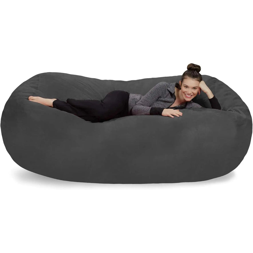 A woman with dark hair in a bun, wearing black pants and a gray zip-up jacket, relaxes on a Bean Bag Lounger - Plush Sofas with Soft Microsuede Cover in charcoal against a white background.