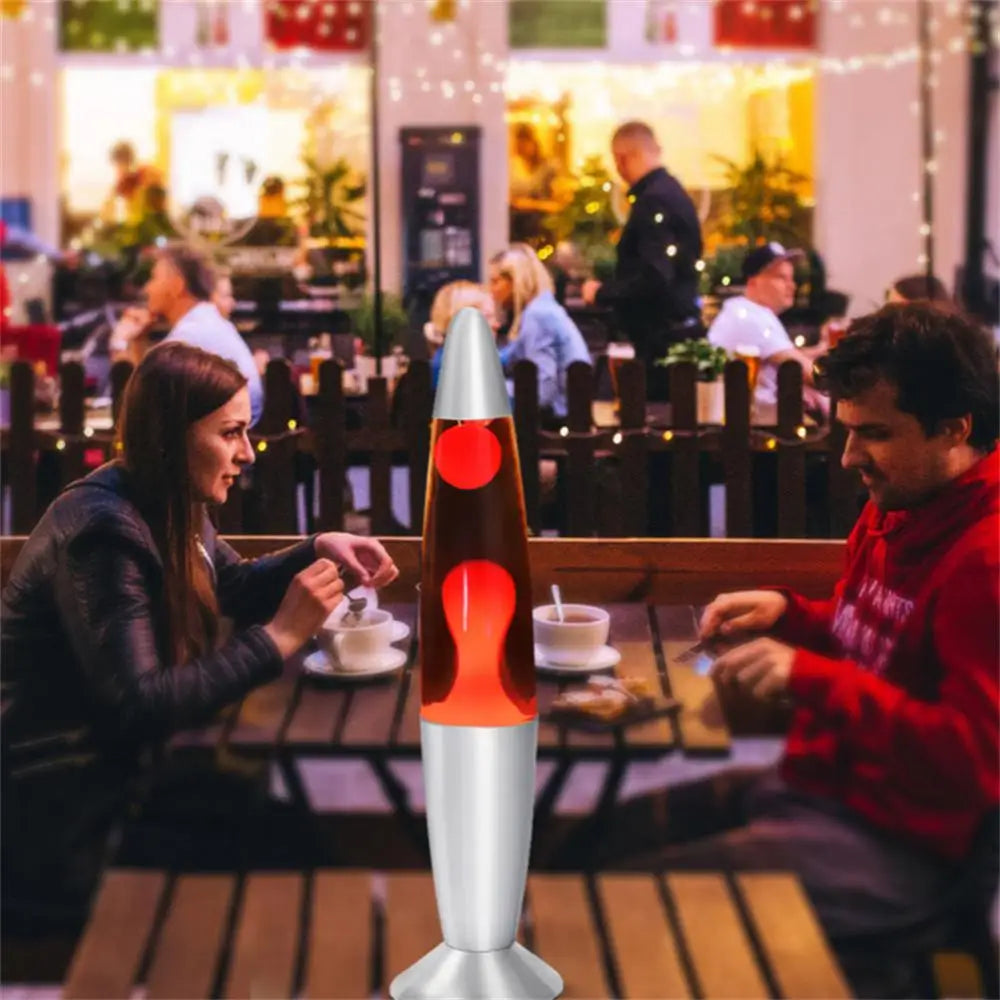 A Mesmerizing Jellyfish Lava LED Night Light lamp amusingly sits on a table between a man and woman enjoying coffee at an outdoor café. The lively scene, enhanced by string lights, mirrors the effervescence of the lamps playful ambiance.