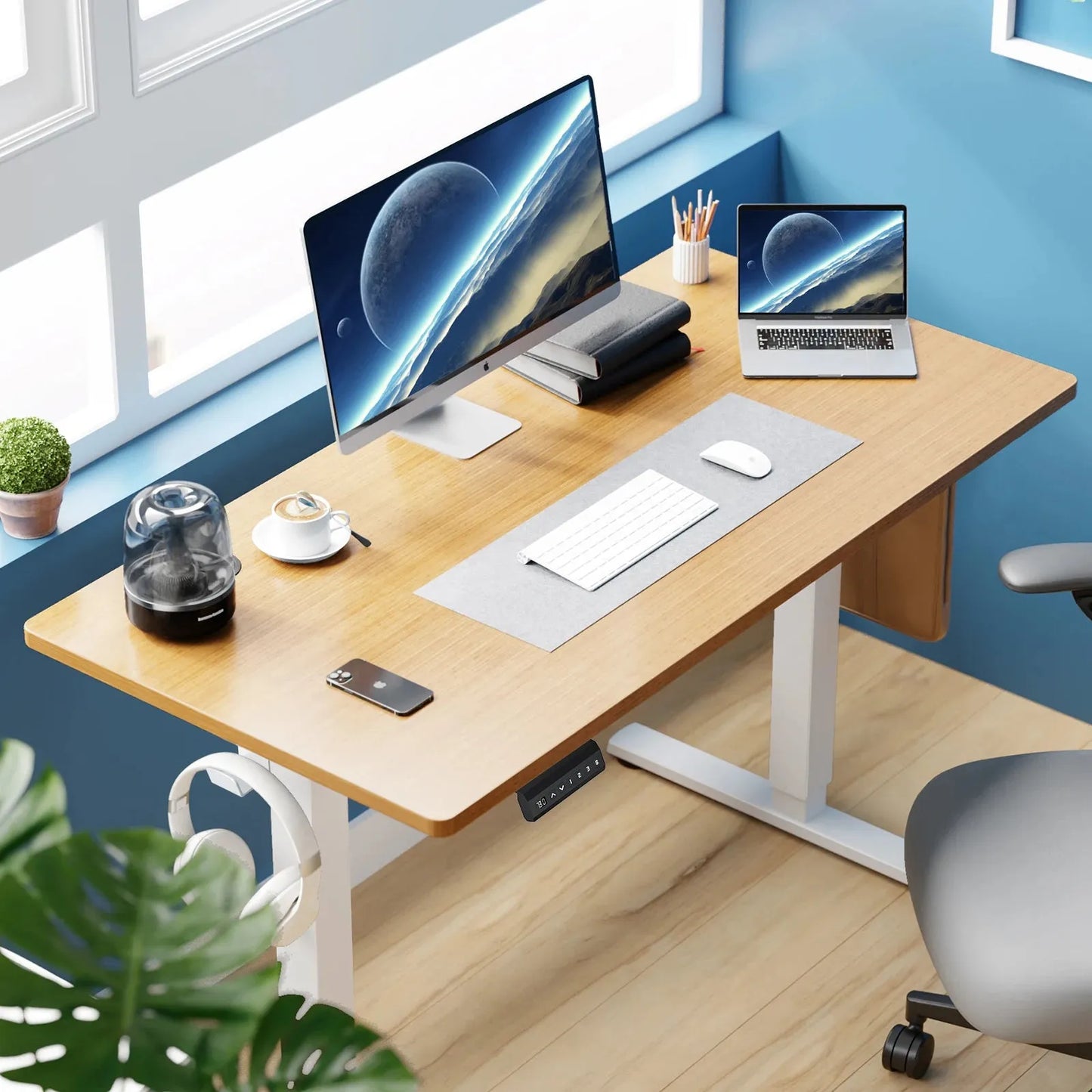The JHK Electric Height Adjustable Standing Desk - Ergonomic 40x24 Inch Sit-Stand Workstation hosts a large monitor and laptop with a space scene, alongside a keyboard, mouse, plant, phone, and compact humidifier. The wooden desk sits against a blue wall illuminated by generous window light.
