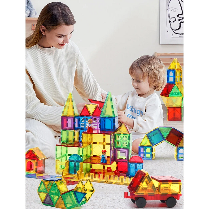 A woman and child enjoy building a castle with the Magnetic Building Blocks with Cars - 3D Clear Educational Construction Set for Kids Ages 3-8, enhancing fine motor skills. A small red toy truck sits near them on a light-colored floor as they smile.