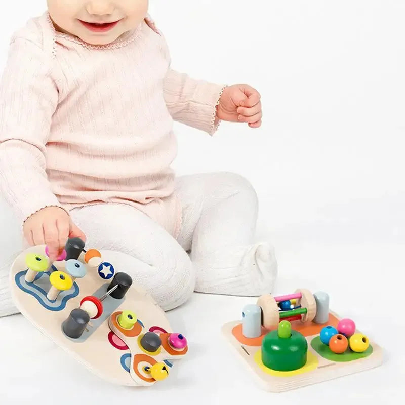 A baby in a light pink outfit and white pants sits on the floor, enjoying educational fun with colorful wooden toys. One toy is an Interactive Wooden Sensory Learning Board for Kids, perfect for developing fine motor skills with movable pieces, vibrant beads, and shapes.